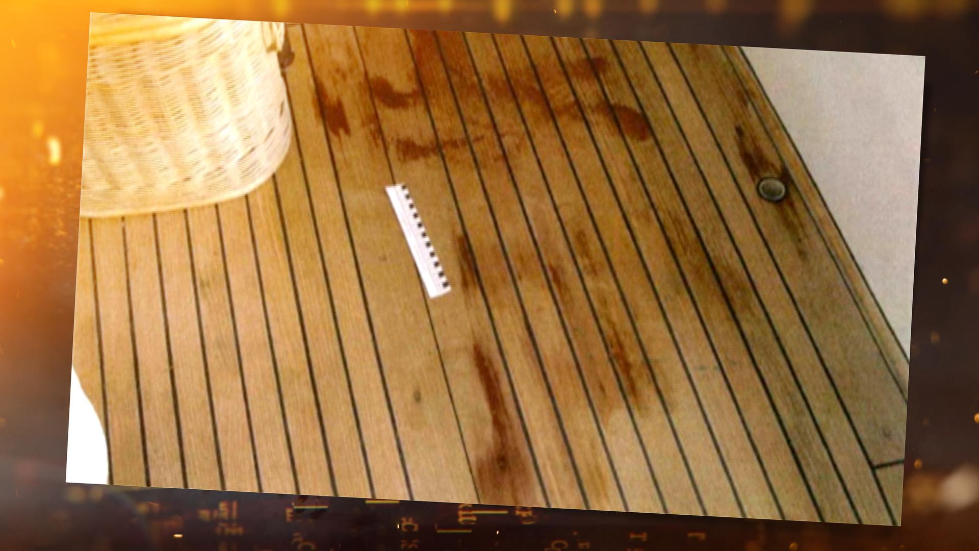 Auf einem braunen Holzboden sind rote Flecken sichtbar. Neben einem beigen Korb liegt ein weiß-schwarzes Lineal, das die Größe der Flecken misst.