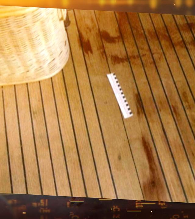 Auf einem braunen Holzboden sind rote Flecken sichtbar. Neben einem beigen Korb liegt ein weiß-schwarzes Lineal, das die Größe der Flecken misst.