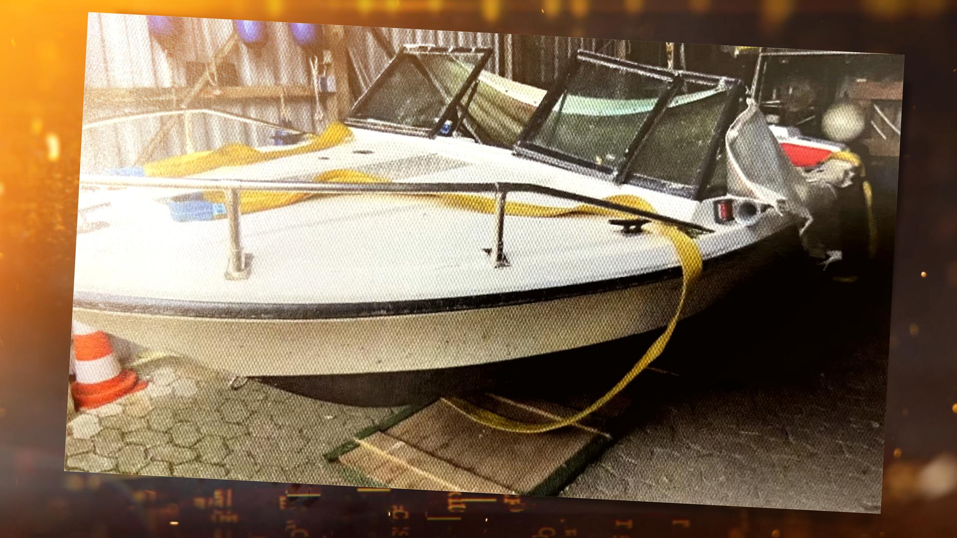 ein weißes Boot in einer Halle mit grauen Metallwänden. Der Bauch des Bootes ist unten schwarz gestrichen und hat oben einen schwarzen länglichen Strich. Darüber ist eine silberne Reling angebracht. Vor dem Führerstand sind drei Fenster mit einem schwarzen Rahmen erkennbar. Das mittlere Fenster ist ausgehebelt und an dem rechten Fenster angelehnt. Der Boden der Halle ist mit grauen Pflastersteinen ausgelegt.