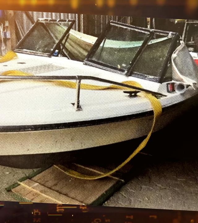 ein weißes Boot in einer Halle mit grauen Metallwänden. Der Bauch des Bootes ist unten schwarz gestrichen und hat oben einen schwarzen länglichen Strich. Darüber ist eine silberne Reling angebracht. Vor dem Führerstand sind drei Fenster mit einem schwarzen Rahmen erkennbar. Das mittlere Fenster ist ausgehebelt und an dem rechten Fenster angelehnt. Der Boden der Halle ist mit grauen Pflastersteinen ausgelegt.