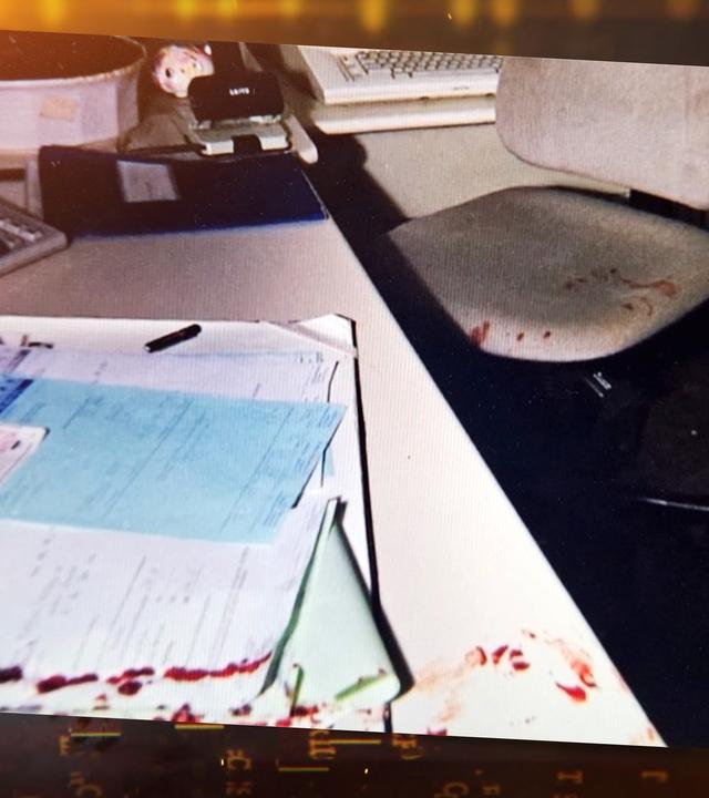 Die Farbfotografie zeigt einen Schreibtisch in einem Büro oder Verwaltungsraum. Im linken Bildbereich liegen mehrere übereinander gestapelte Blätter und Formulare auf dem hellen Tisch – teils weiß, teils blau. Einige der Dokumente sind am unteren Rand deutlich mit blutigen Abdrücken oder Spritzern beschmutzt. Ein schwarzer Kugelschreiber liegt zwischen den Papieren.
