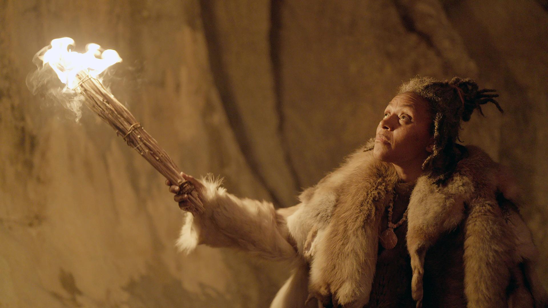 Ein Frau mit einer Fackel leuchtet eine Felswand an. Sie ist in Tierfelle gekleidet und trägt eine Muschel als Halskette.