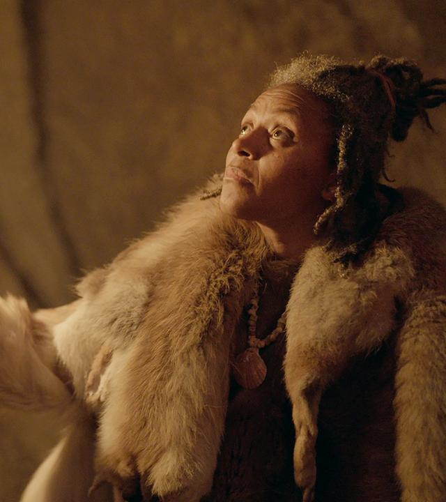 Ein Frau mit einer Fackel leuchtet eine Felswand an. Sie ist in Tierfelle gekleidet und trägt eine Muschel als Halskette.