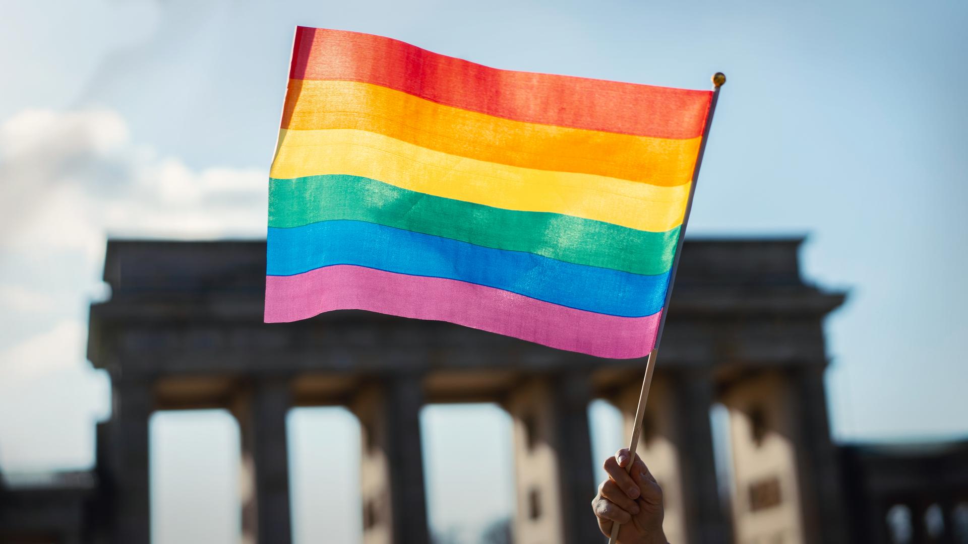 EIne Hand schwenkt eine Regenbogenfahne vor dem Brandenburger Tor.