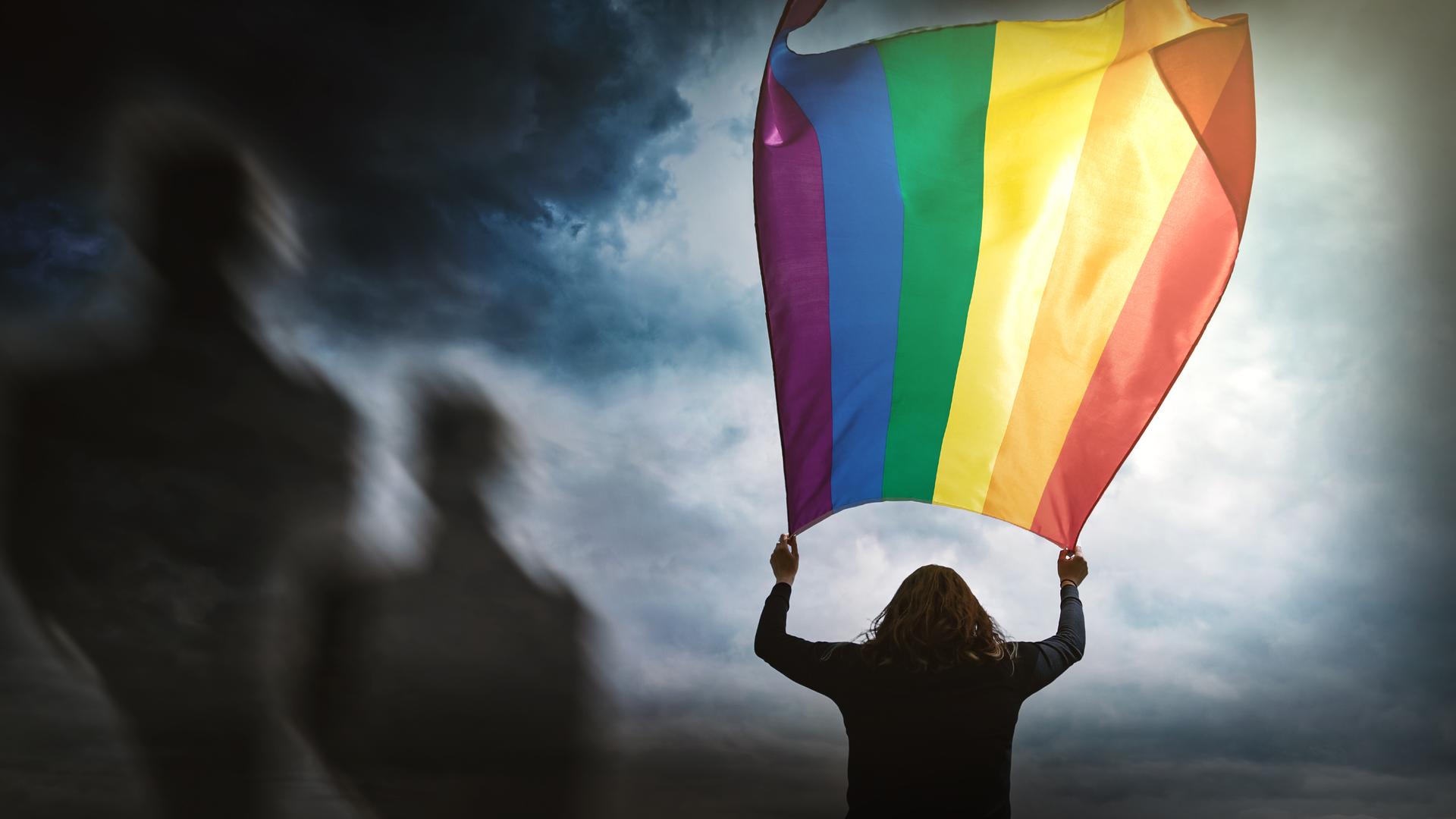 Silhouette einer Person, die mit ausgestreckten Armen eine große Regenbogenfahne über sich hält. Im Hintegrund sind schemenhaft weitere Personen zu erkennen.