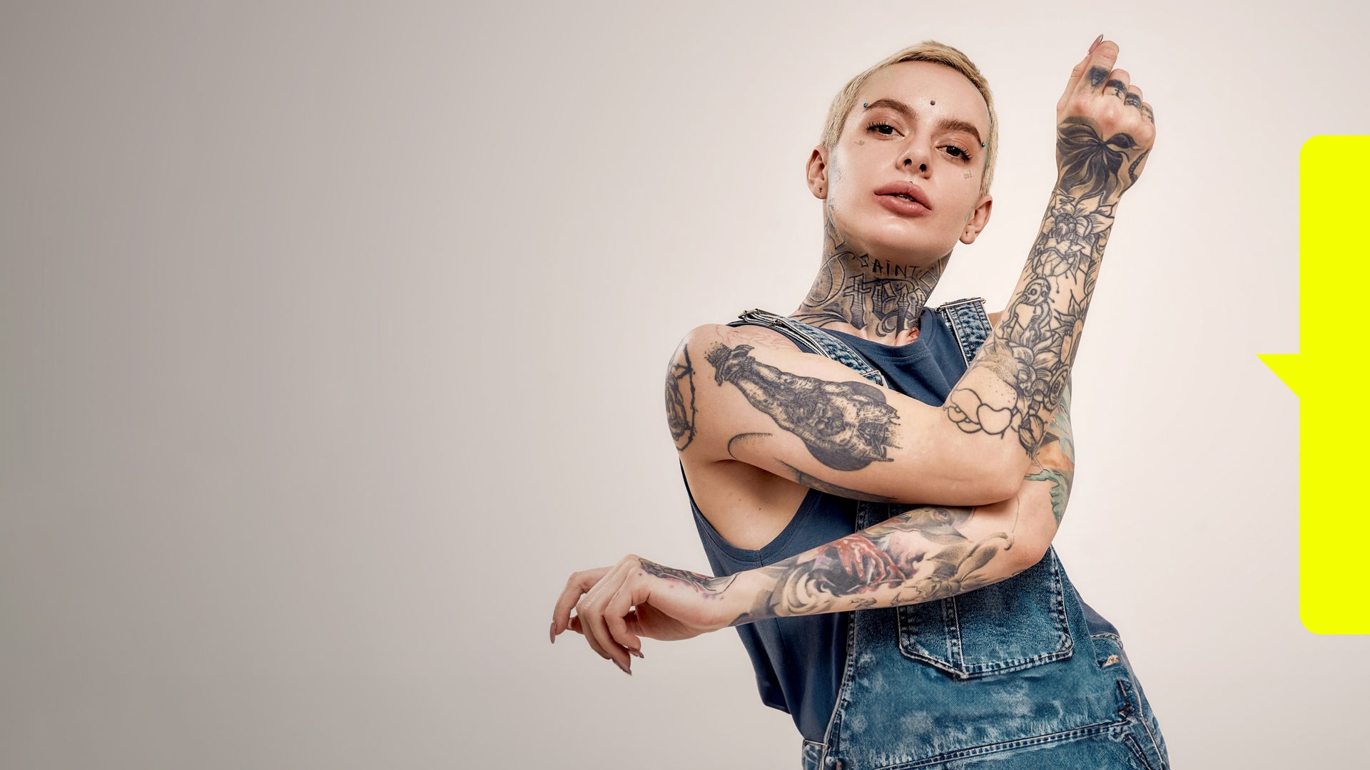 Eine junge Frau mit vielen sichtbaren Tätowierungen an Armen und Hals sowie mehreren Piercings im Gesicht.