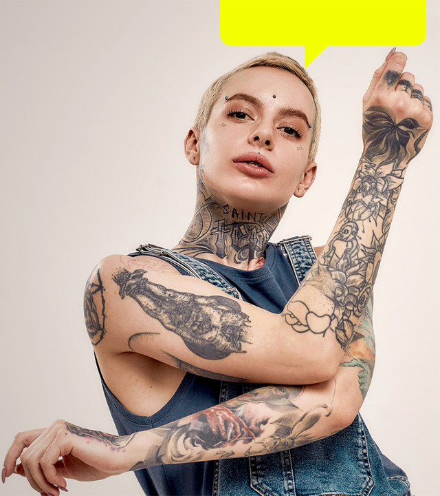 Eine junge Frau mit vielen sichtbaren Tätowierungen an Armen und Hals sowie mehreren Piercings im Gesicht.