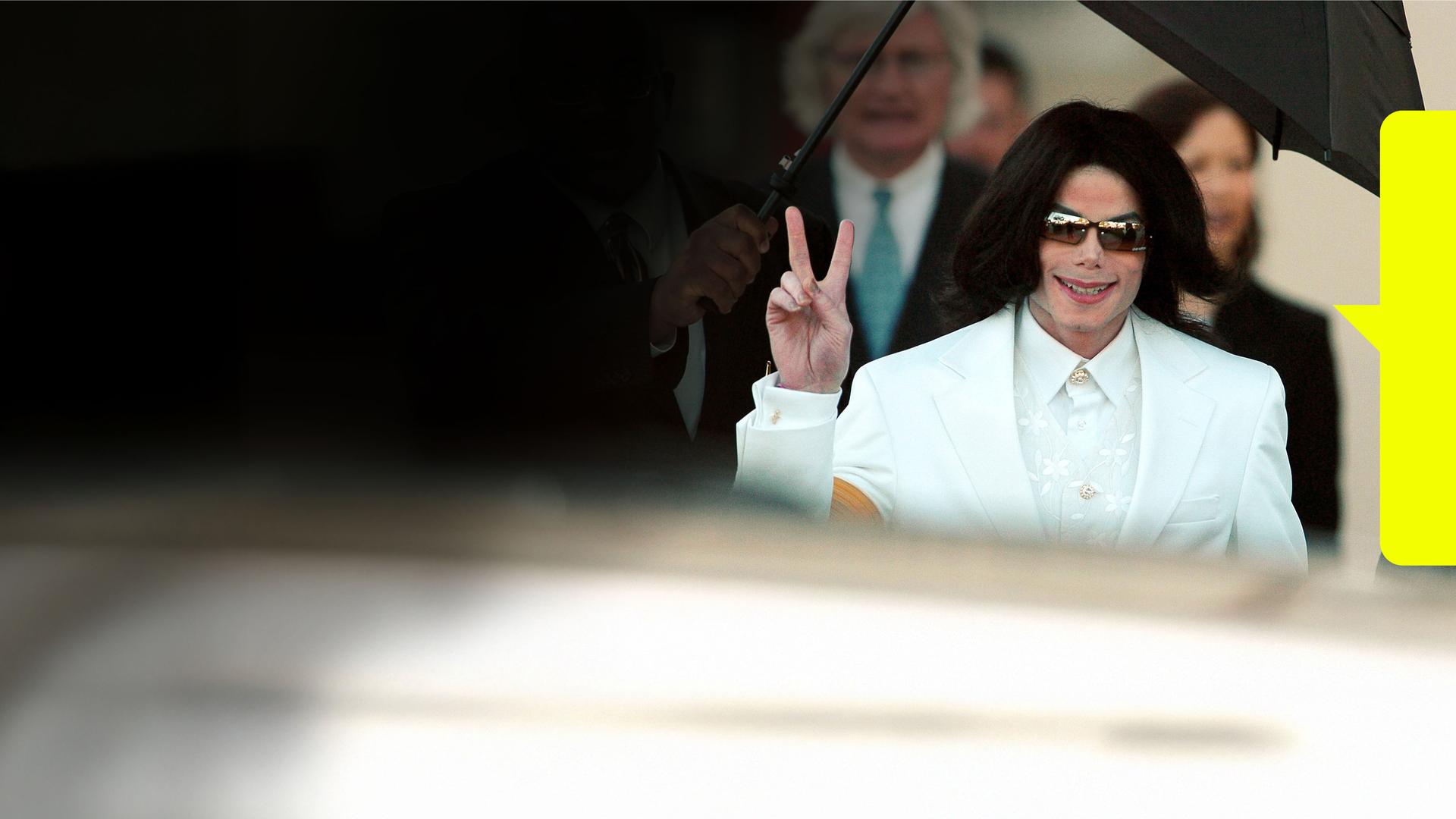 Micheal Jackson in weißem Anzug auf dem Weg zum Gericht, zeigt mit der rechten hand das Peace-Zeichen