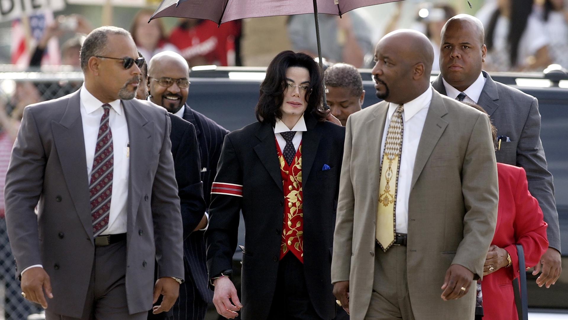 Michael Jackson ist auf dem Weg zum Gericht in Santa Maria im Jahr 2005. Der Popstar ist von seinem Team umgeben. Er trägt einen schwarzen Anzug mit einer auffälligen roten Weste und einer roten Armbinde. Eine Begleiterin hält einen Schirm über ihn.