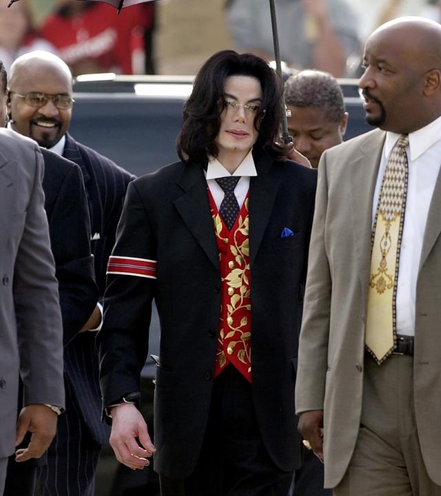 Michael Jackson ist auf dem Weg zum Gericht in Santa Maria im Jahr 2005. Der Popstar ist von seinem Team umgeben. Er trägt einen schwarzen Anzug mit einer auffälligen roten Weste und einer roten Armbinde. Eine Begleiterin hält einen Schirm über ihn.