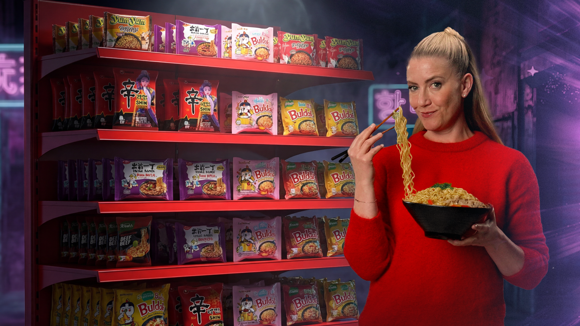 Pia Osterhaus steht mit einer Nudel-Bowl und Essstäbchen in der Han vor einem Regal voller Asia-Tüttensuppen. Mit den Essstäbchen hebt sie Nudeln aus der Bowl Richtung Mund und schaut in die Kamera.