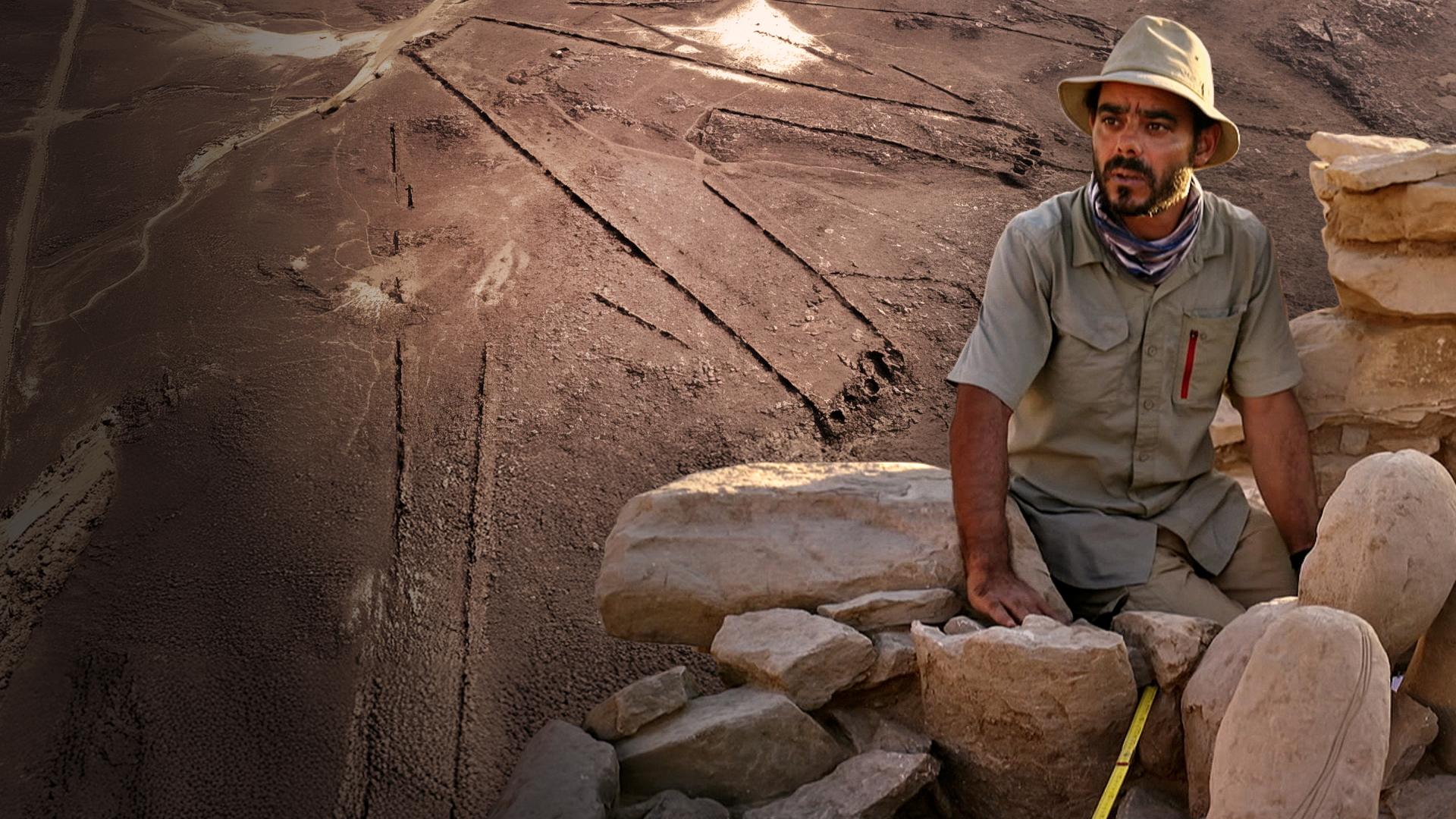 Vorne sitzt ein Forscher bei der Untersuchung einer Ruine nahe eines "Wüstendrachen" auf Steinen. Ein solcher "Wüstendrache" ist im Hintergrund als Luftaufnahme zu sehen ist.