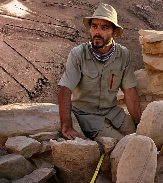 Vorne sitzt ein Forscher bei der Untersuchung einer Ruine nahe eines "Wüstendrachen" auf Steinen. Ein solcher "Wüstendrache" ist im Hintergrund als Luftaufnahme zu sehen ist.