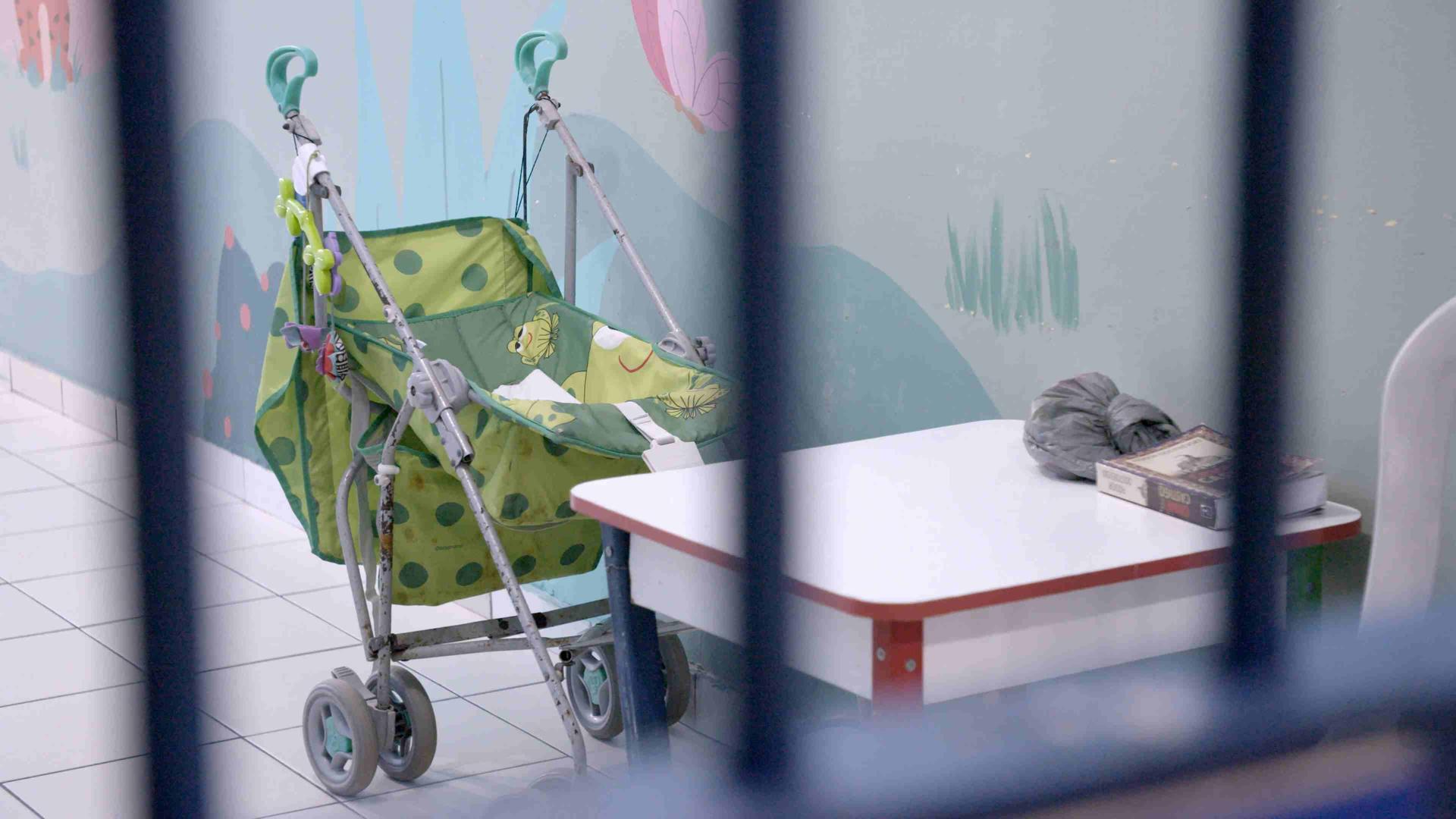 Zwischen unscharfen Gitterstäben ist ein hellgrüner Buggy mit Froschmotiv zu sehen. Er parkt vor einer farbenfroh bemalten Wand. Vor dem Buggy steht ein kleiner Kindertisch.