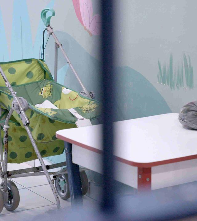 Zwischen unscharfen Gitterstäben ist ein hellgrüner Buggy mit Froschmotiv zu sehen. Er parkt vor einer farbenfroh bemalten Wand. Vor dem Buggy steht ein kleiner Kindertisch.