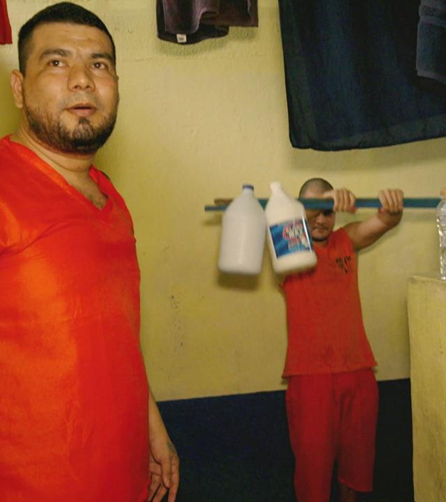 Zwei Häftlinge in roten T-Shirts stehen in einem Gefängnisraum. Einer von ihnen stemmt eine selbst gebaute Hantelstange, an deren Enden mehrere Wasserkanister als Gewicht befestigt sind.