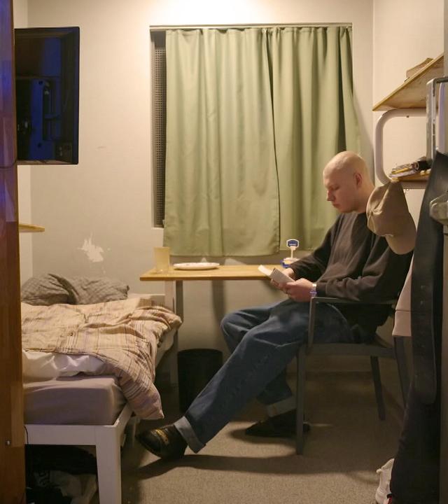 EIn Häftling sitzt alleine auf einem Stuhl in siener kleinen Gefängniszelle. Neben ihm stehen ein Bett und ein Tisch, rechts ist die schwere Stahltür geöffnet.