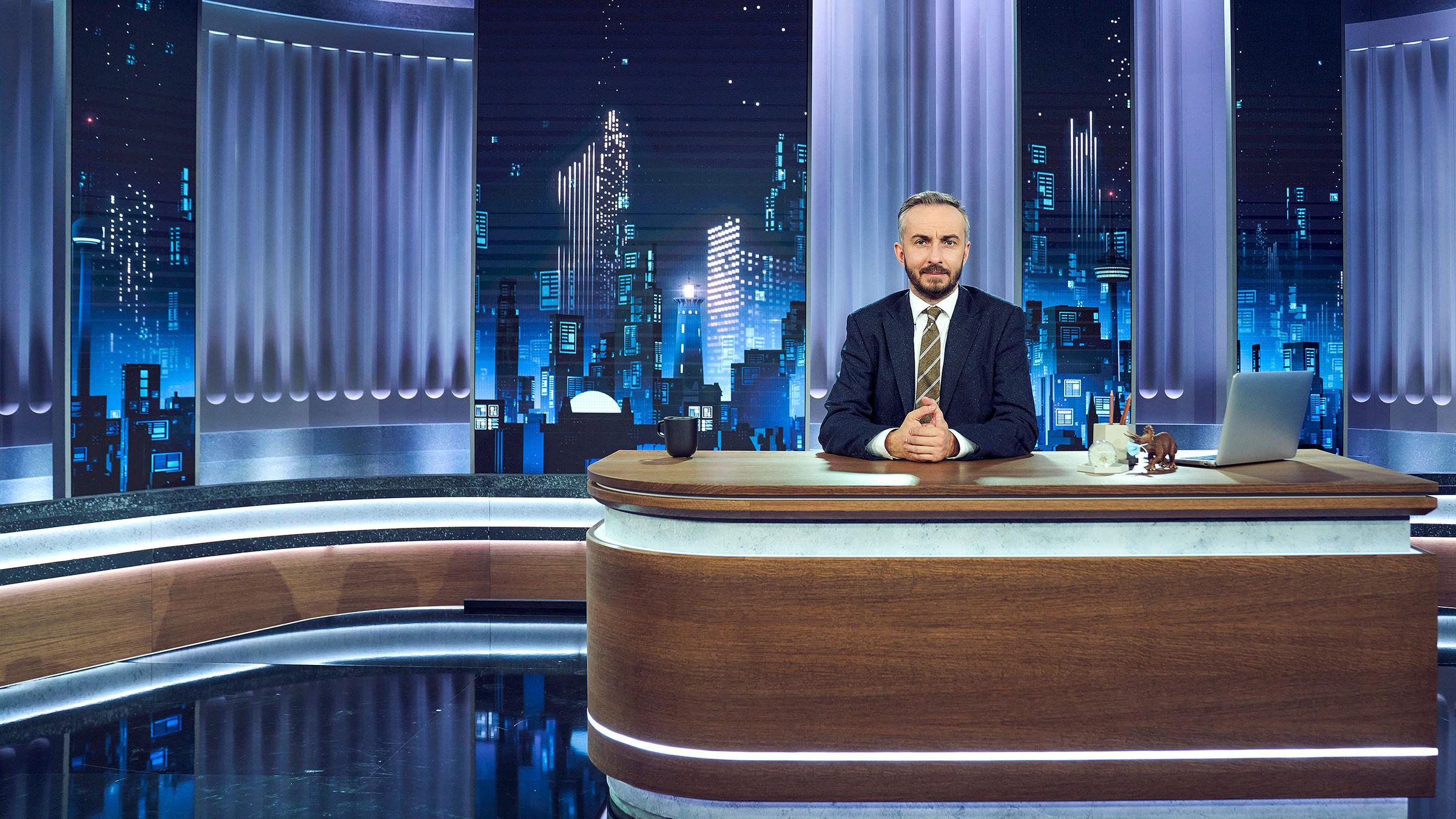 Moderator Jan Böhmermann sitzt am Schreibtisch des neuen ZDF Magazin Royale Studios. Die Hände gefaltet. Hinter ihm eine Late-Night-Skyline.