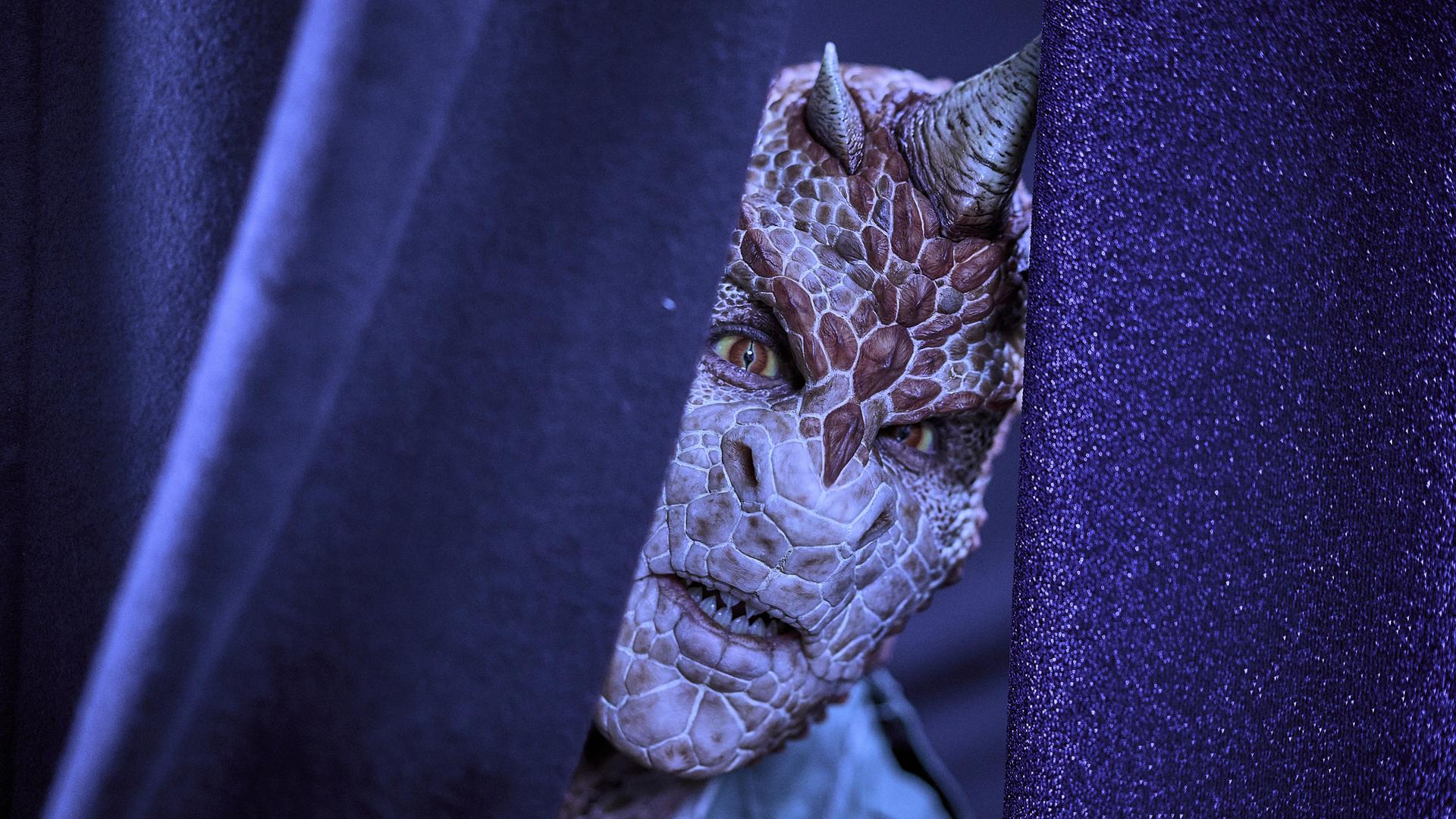 Ein Mensch mit Echsen-Gesicht schaut durch einen leicht aufgeschobenen Bühnenvorhang