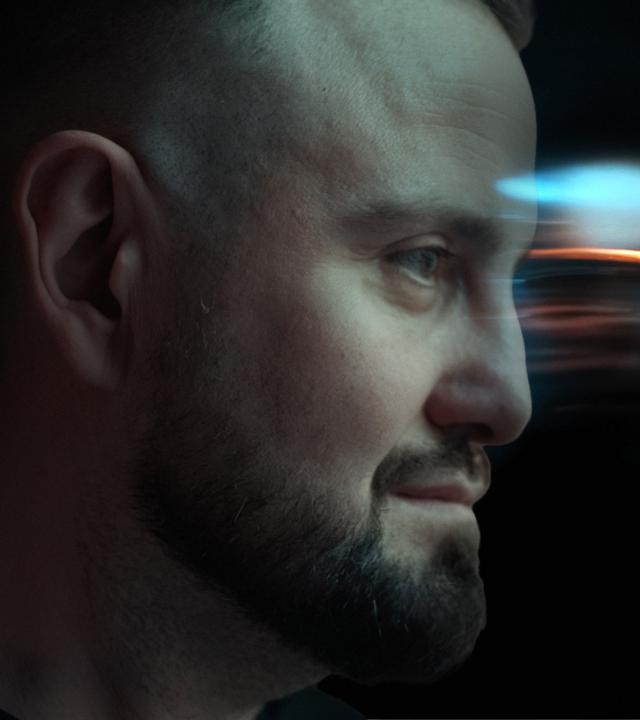 Collage mit zwei Gesichtern. Das eine ist im Profil nach rechts schauend - Hacker "BadB", ein Mann mit Glatze und Bart. Das andere Gesicht ist eher undeutlich als Profil nach links schauend im Hintergrund zu sehen.