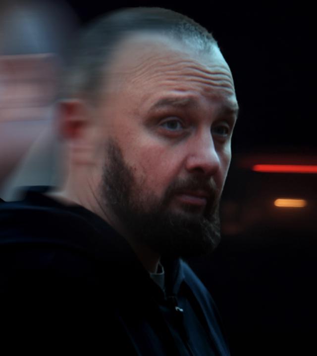 Eine Kollage aus zwei Gesichtern. Das rechte Gesicht zeigt einen Mann mit Vollbart, Blickrichtung nach vorne rechts. In Hintergrund ist Putins Gesicht zu sehen, im Profil verschwommen nach links schauend.