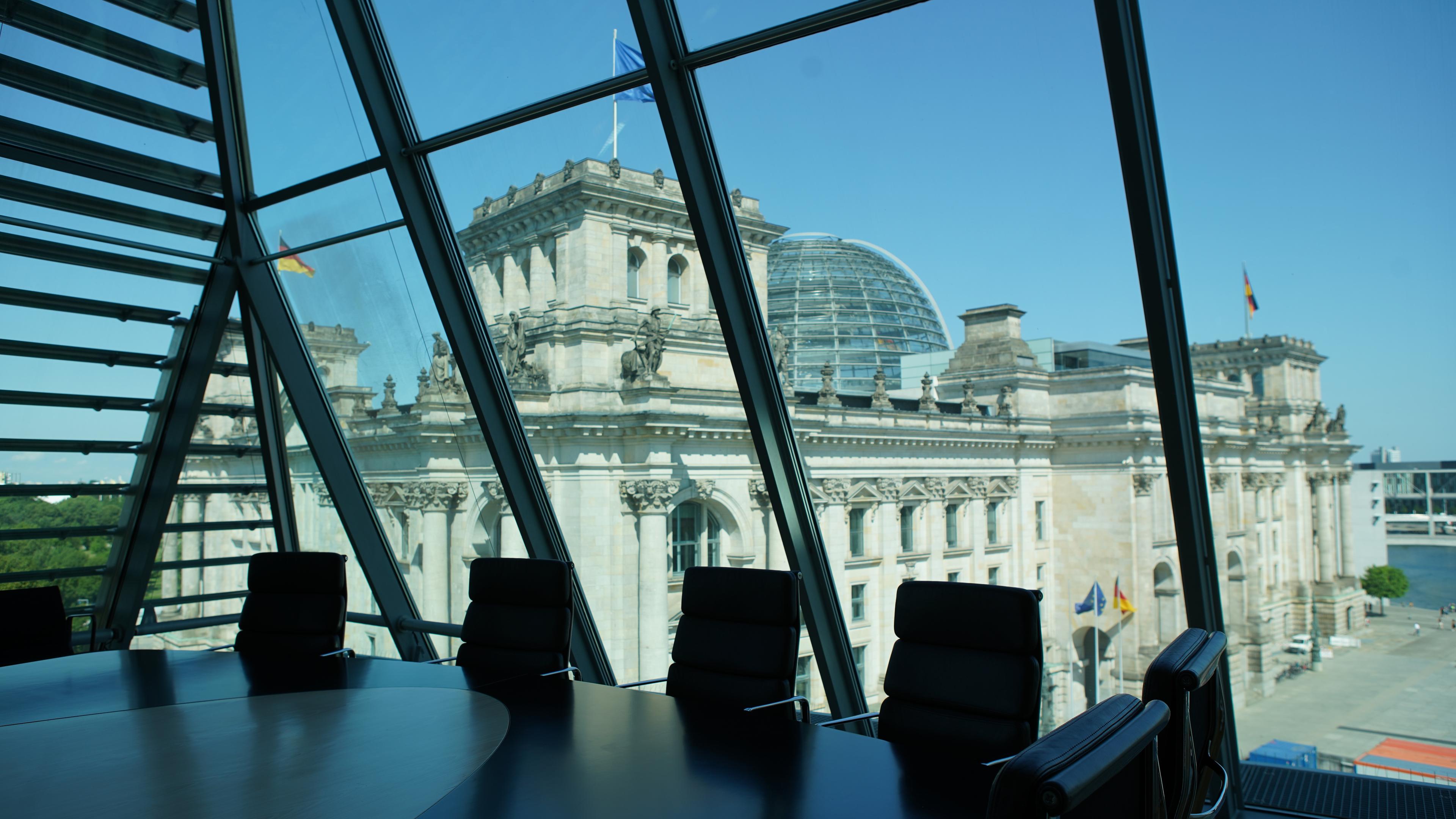 Im Vordergrund ein Tisch mit Stühlen. Im Hintergrund sieht man den Reichstag durch eine Fensterfront.