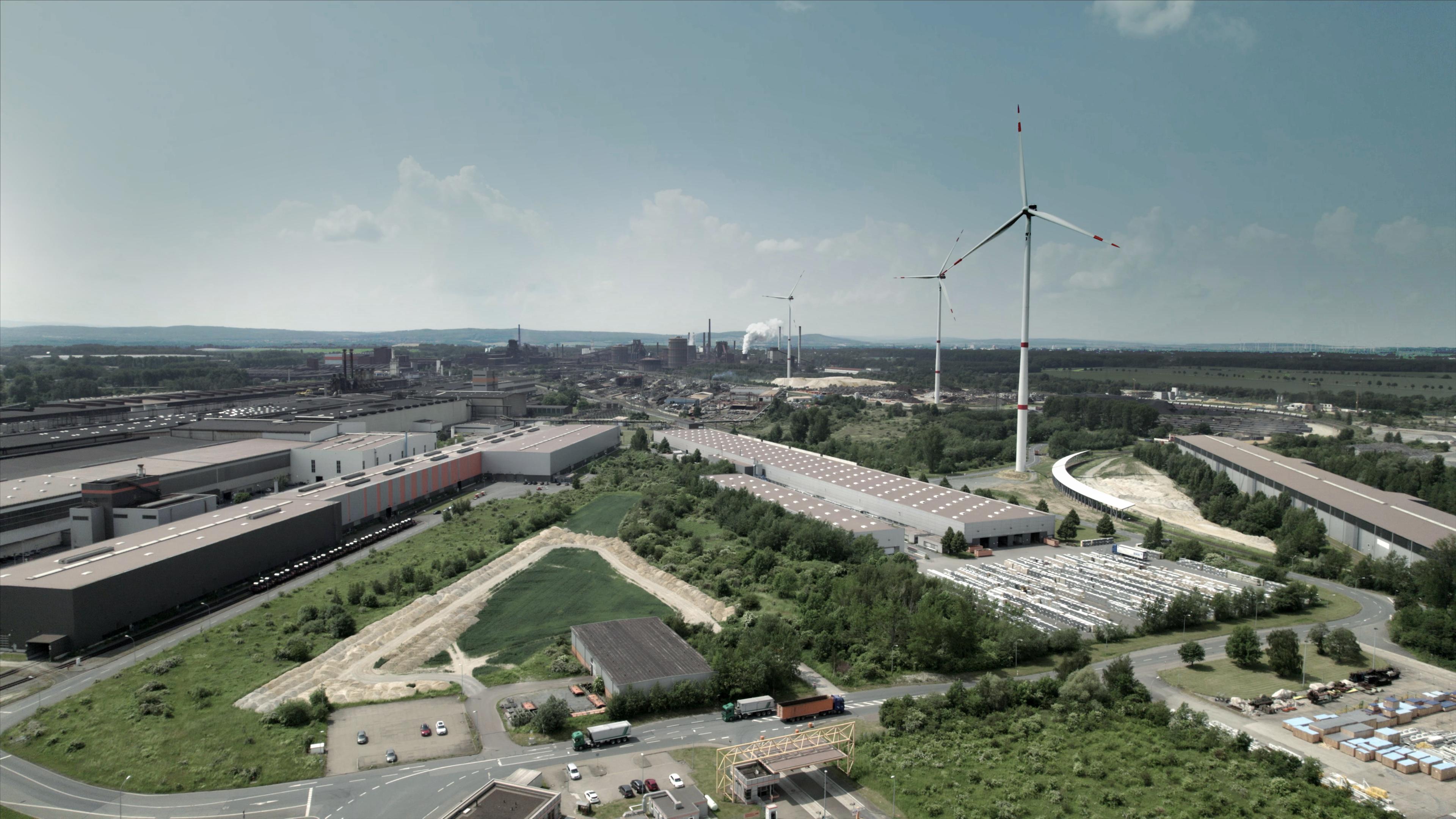 Werksgelände des Stahlwerks Salzgitter mit Gebäuden, Straßen, Parkplätzen, Grünflächen und Windrädern.