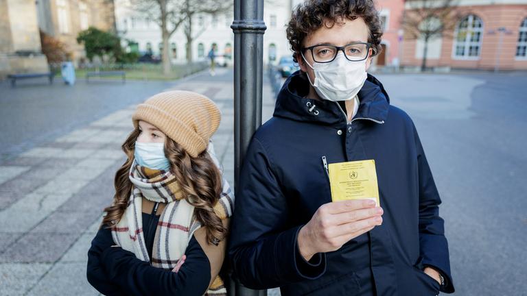 Junge Frau mit Mantel, Mütze, Schal, Mundschutz und verschränkten Armen und junger Mann mit Brille, blauem Kapuzenmantel, Mundschutz und hochgehaltenem gelbem Impfpass, stehen voneinander abgewandt an einer schwarzen Straßenlaterne.