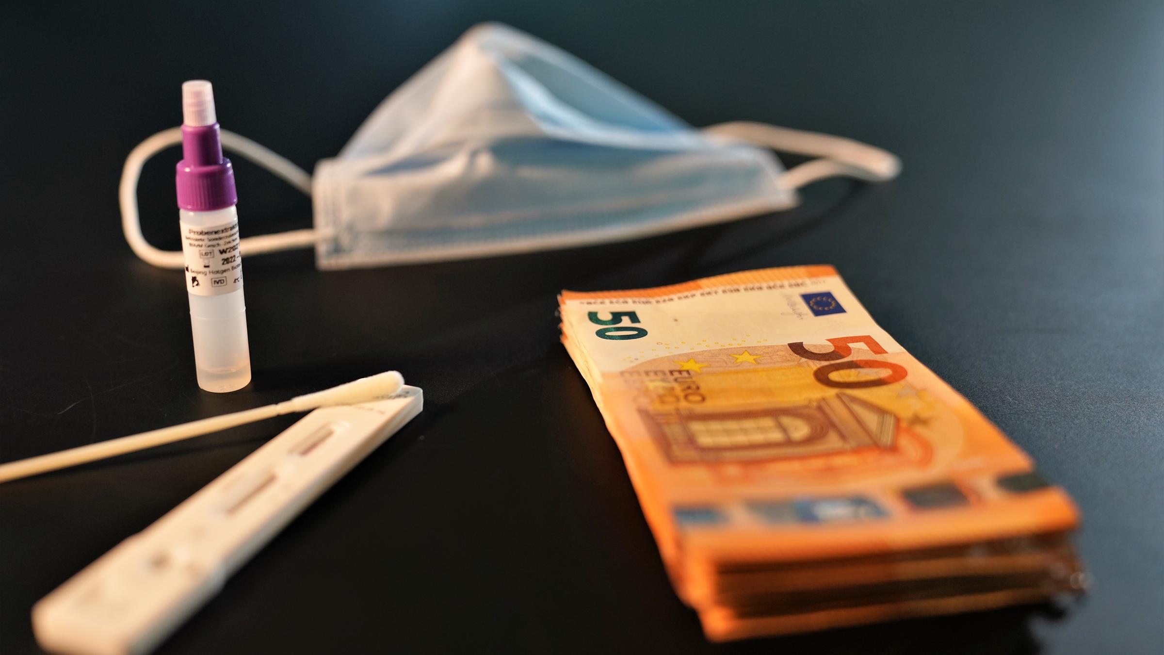 ZDFzoom: Kassensturz - Wer zahlt die Corona-Kosten: Schnelltest-Utensilien und Maske neben einem Bündel 50-Euro-Scheine.