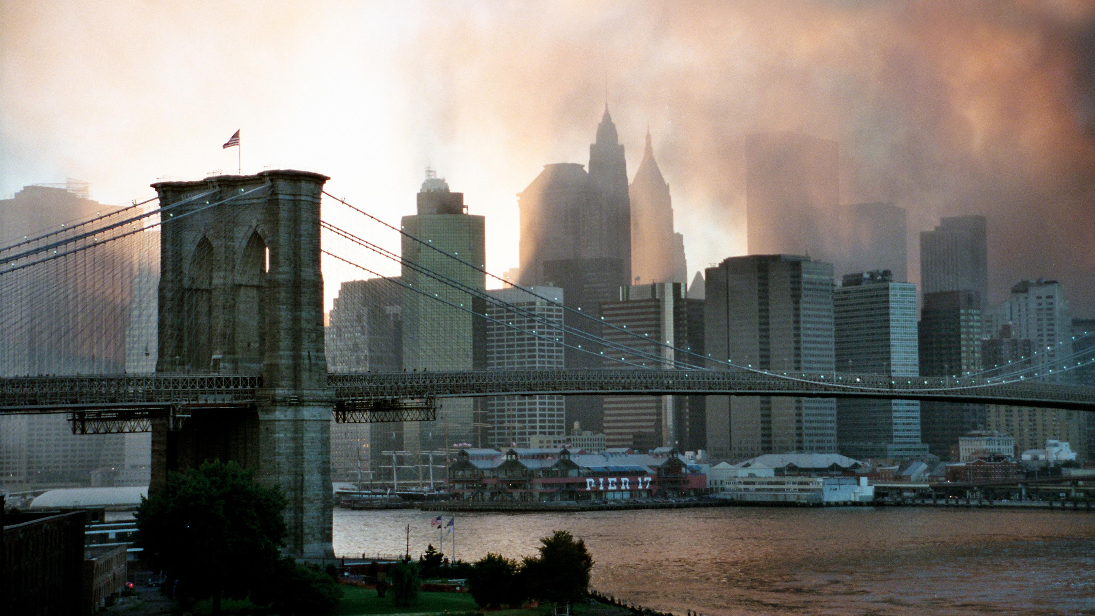 "ZDFzoom: 20 Jahre 9/11 - Ein Tag im September": Brooklyn-Bridge im Vordergrund, im Hintergrund Skyline von Manhattan im Sonnenuntergang und umhüllt von Rauch.