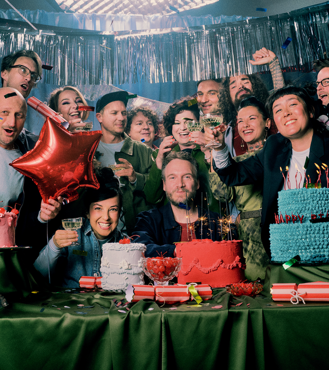Die Musikerinnen und Musiker des "Rundfunk-Tanzorchesters Ehrenfeld" sitzen oder stehen hinter einem mit bunten Torten gedeckten Tisch und stoßen mit Sektgläsern an.