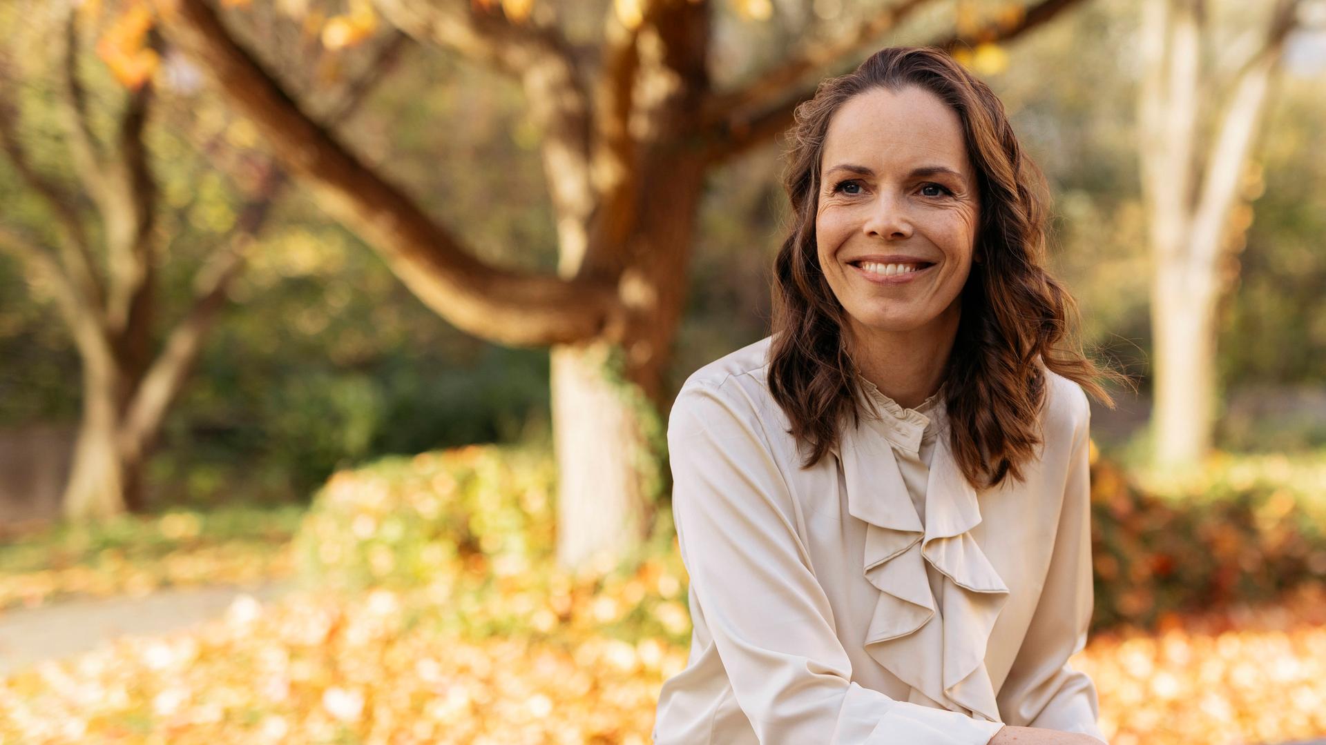 Juliane sitzt auf einer Bank und strahlt lächelnd in die Kamera. Im Hintergrund sind herbstliche Bäume zu sehen, die ihre Blätter verlieren. 