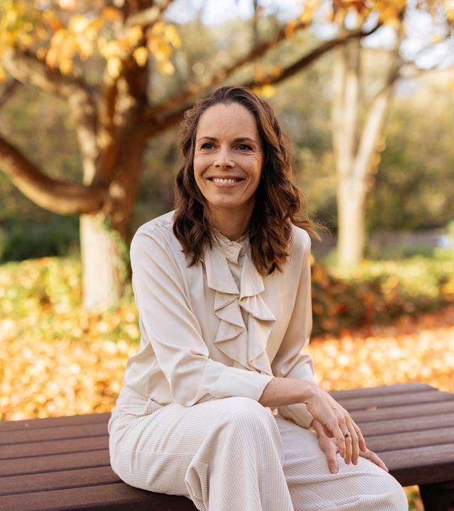Juliane sitzt auf einer Bank und strahlt lächelnd in die Kamera. Im Hintergrund sind herbstliche Bäume zu sehen, die ihre Blätter verlieren. 