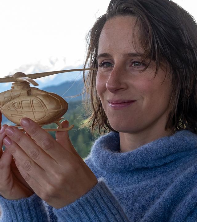 Unter einem Verschlag mit Blick auf eine Berglandschaft steht Maike (Amelie Kiefer) und hält lächelnd einen Modellhubschrauber aus Holz in den Händen.