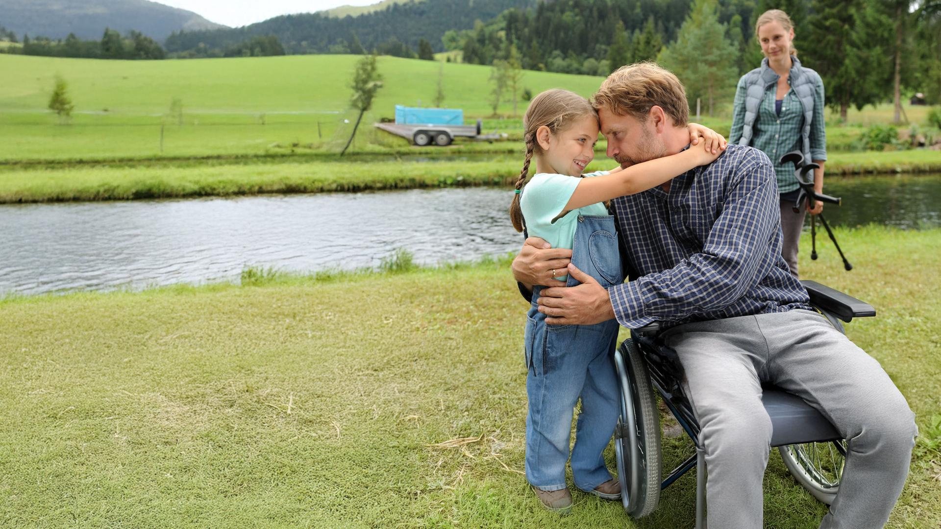 "Zeit für Frühling": Die kleine Lilly (Helena Pieske) umarmt ihren Vater Franz Holzner (Janek Rieke) am Forellen-Teich. Sandra Holzner (Picco von Groote) beobachtet die beiden aus dem Hintergrund.