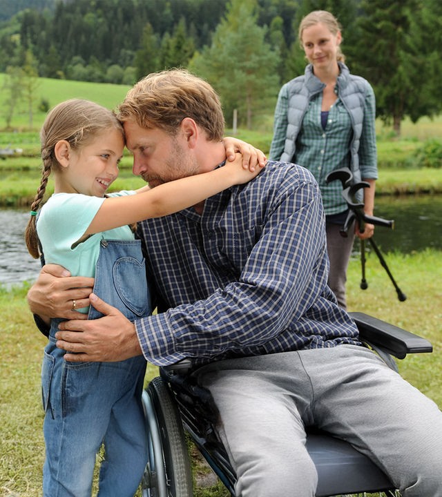 "Zeit für Frühling": Die kleine Lilly (Helena Pieske) umarmt ihren Vater Franz Holzner (Janek Rieke) am Forellen-Teich. Sandra Holzner (Picco von Groote) beobachtet die beiden aus dem Hintergrund.