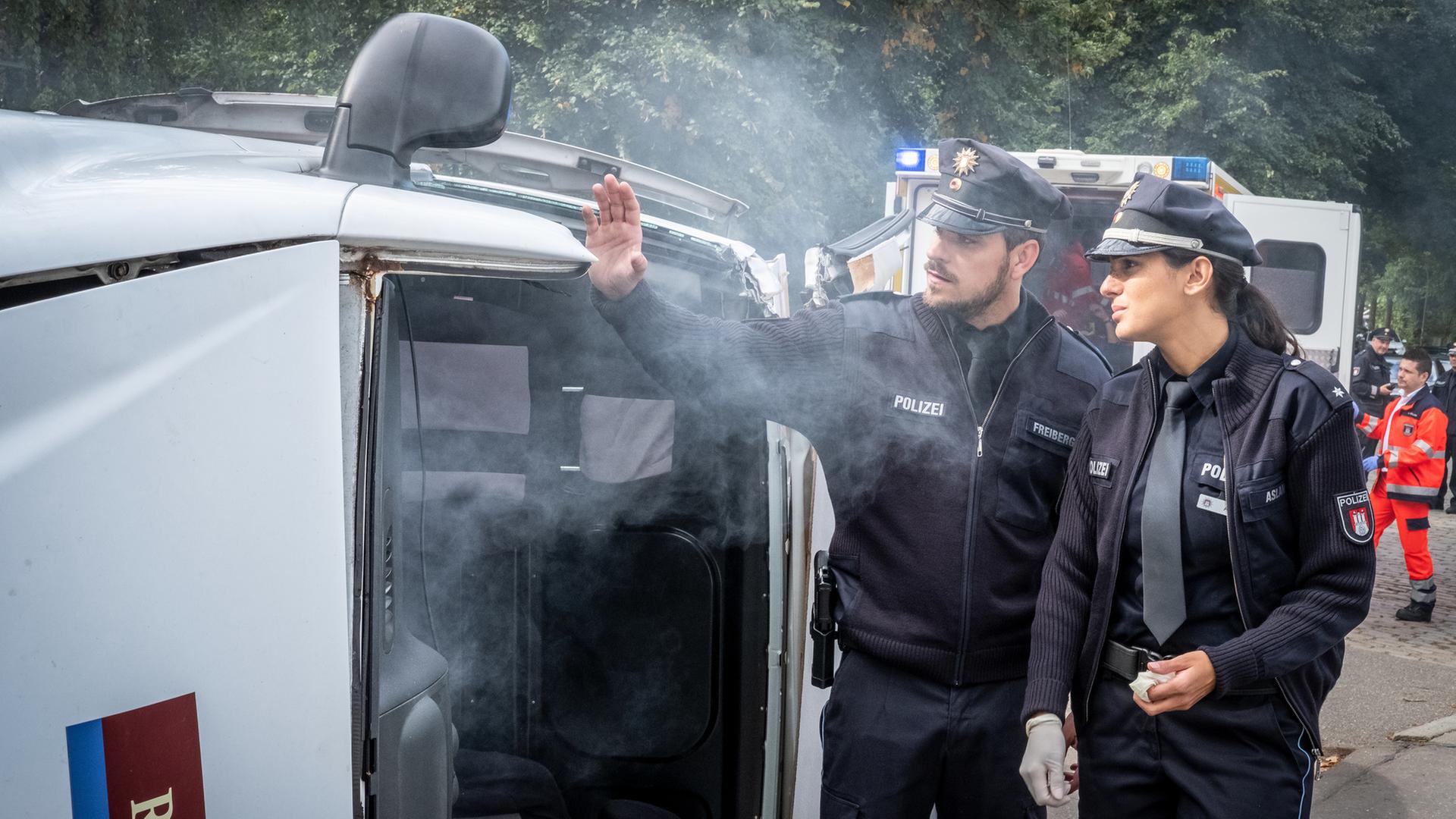 "Notruf Hafenkante - Zeit ist Geld": Kris Freiberg (Marc Barthel) und Pinar Aslan (Aybi Era) stehen neben einem umgekippten Kleintransporter. Qualm steigt aus dem Fahrzeug.