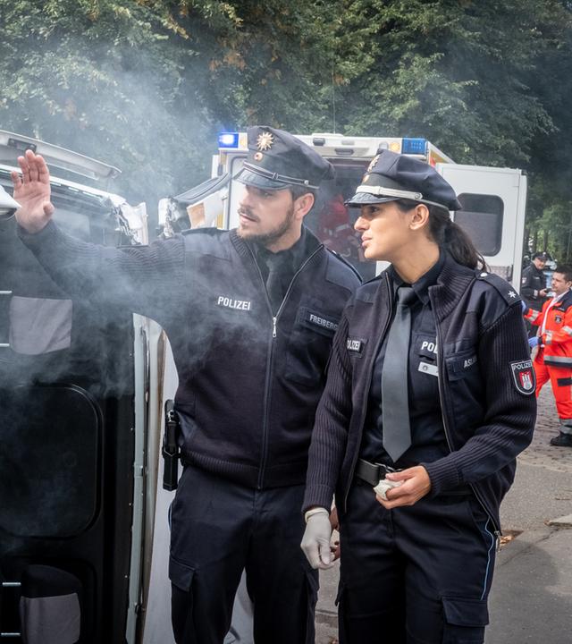 "Notruf Hafenkante - Zeit ist Geld": Kris Freiberg (Marc Barthel) und Pinar Aslan (Aybi Era) stehen neben einem umgekippten Kleintransporter. Qualm steigt aus dem Fahrzeug.
