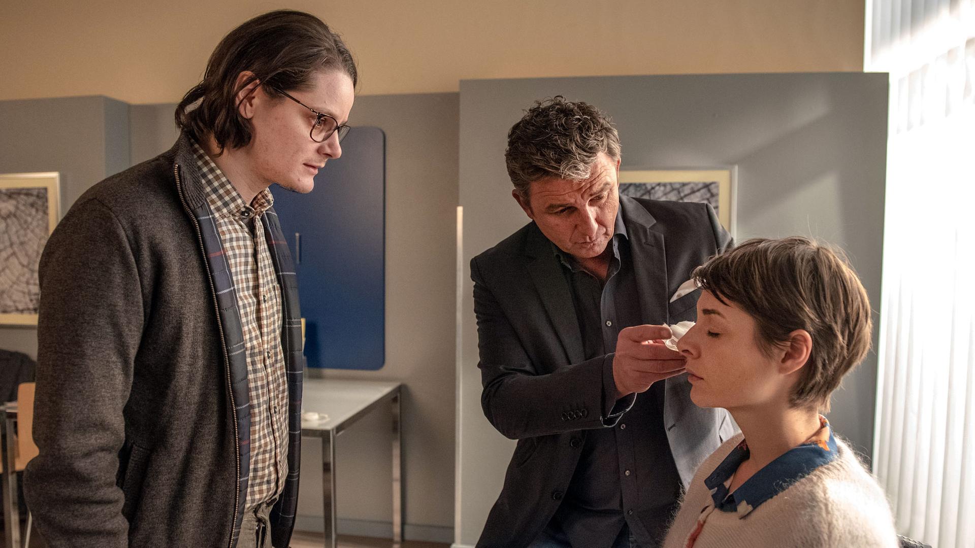 "Der Bergdoktor: Zeitenwende": Im Behandlungszimmer sitzt Jana (Katharina Nesytowa) auf dem Stuhl während Dr. Gruber (Hans Sigl) ihr den Verband von den Augen nimmt. Ihr Freund Anton (Johannes Franke) steht ihr gegenüber.