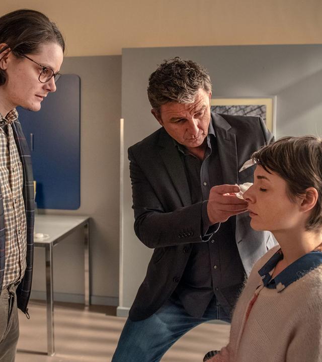 "Der Bergdoktor: Zeitenwende": Im Behandlungszimmer sitzt Jana (Katharina Nesytowa) auf dem Stuhl während Dr. Gruber (Hans Sigl) ihr den Verband von den Augen nimmt. Ihr Freund Anton (Johannes Franke) steht ihr gegenüber.