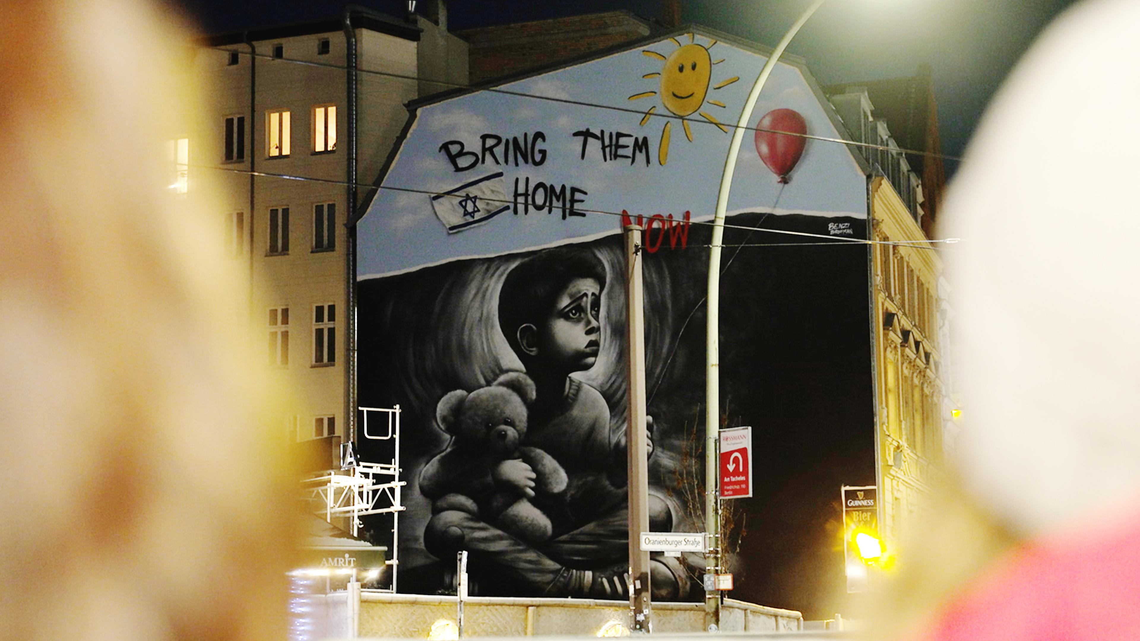 Graffiti an Hauswand zeigt Kind, das Teddy umschlungen hat. Darüber ein Schriftzug: Bring them Home. Und eine Israel-Flagge.