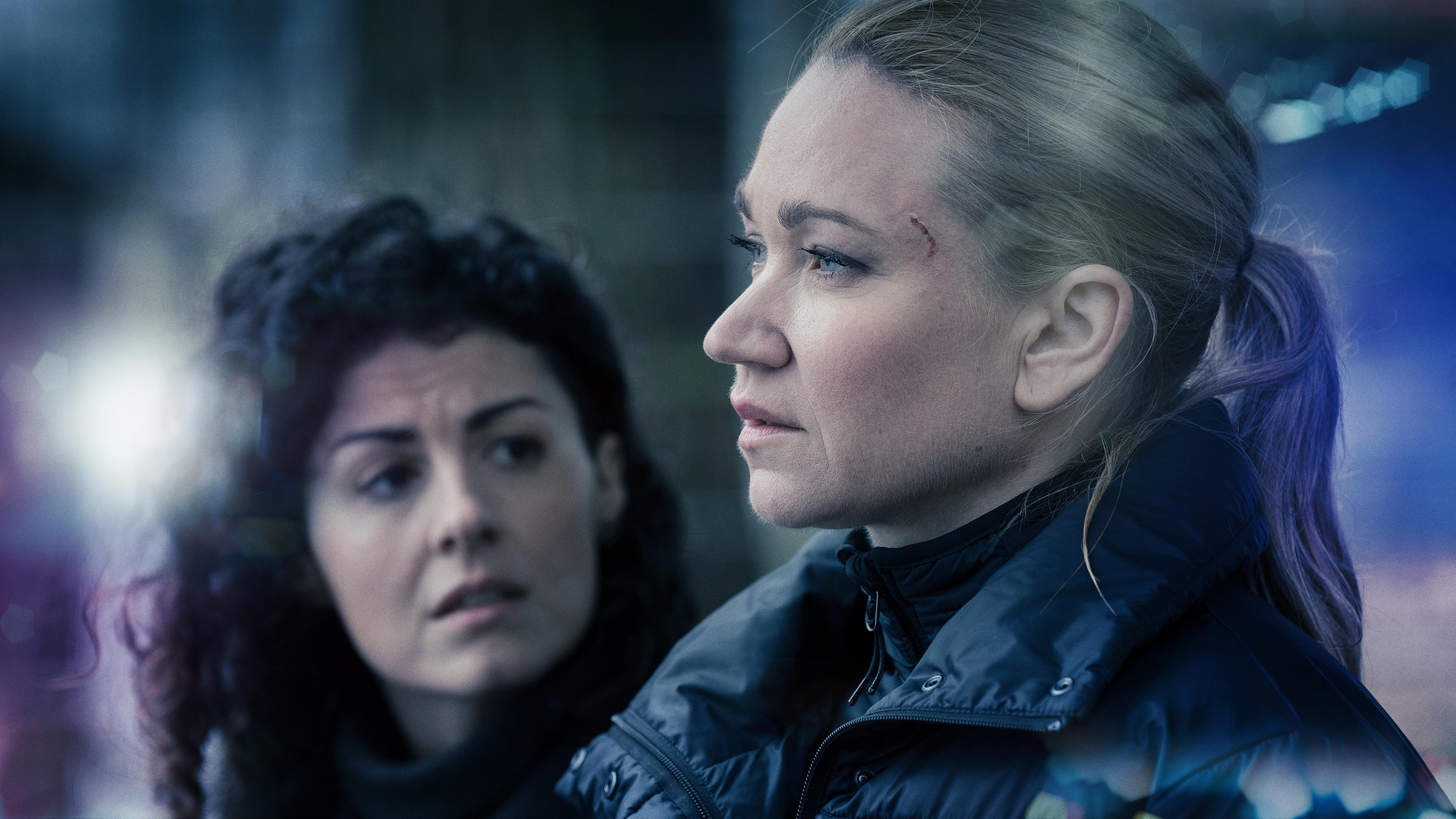 Sarah Kohr (Lisa Marie Potthoff) im Profil mit einer Schramme an der Augenbraue und einer schwarzen Winterjacke. Im Hintergrund wendet sich Charlotte May (Liza Tzschirner), eine Frau mit schwarzen Locken, zu ihr. 