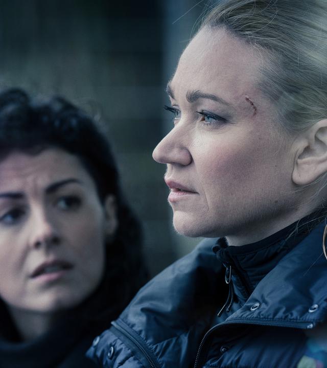 Sarah Kohr (Lisa Marie Potthoff) im Profil mit einer Schramme an der Augenbraue und einer schwarzen Winterjacke. Im Hintergrund wendet sich Charlotte May (Liza Tzschirner), eine Frau mit schwarzen Locken, zu ihr. 