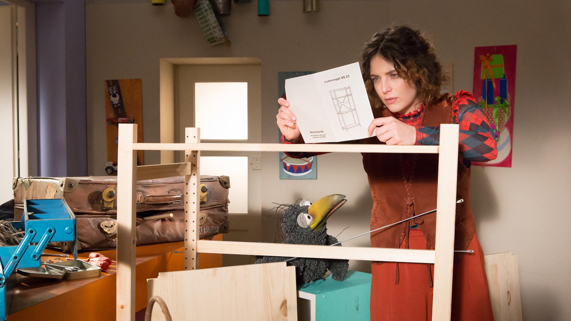 Koffer und Rudi schauen zu Siebenstein, die an einem halb aufgebauten Regal steht und nachdenklich auf eine Bauanleitung schaut.