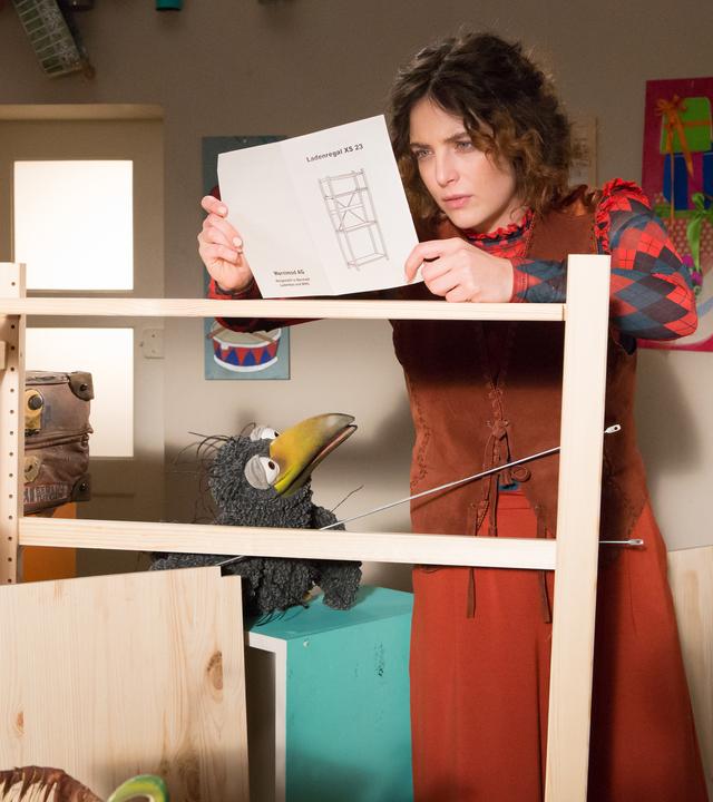 Koffer und Rudi schauen zu Siebenstein, die an einem halb aufgebauten Regal steht und nachdenklich auf eine Bauanleitung schaut.