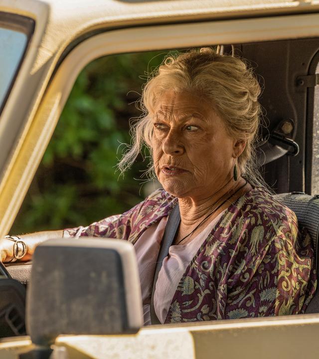 Dans Oma Mitch sitzt in einem gelben Auto hinter dem Steuer.