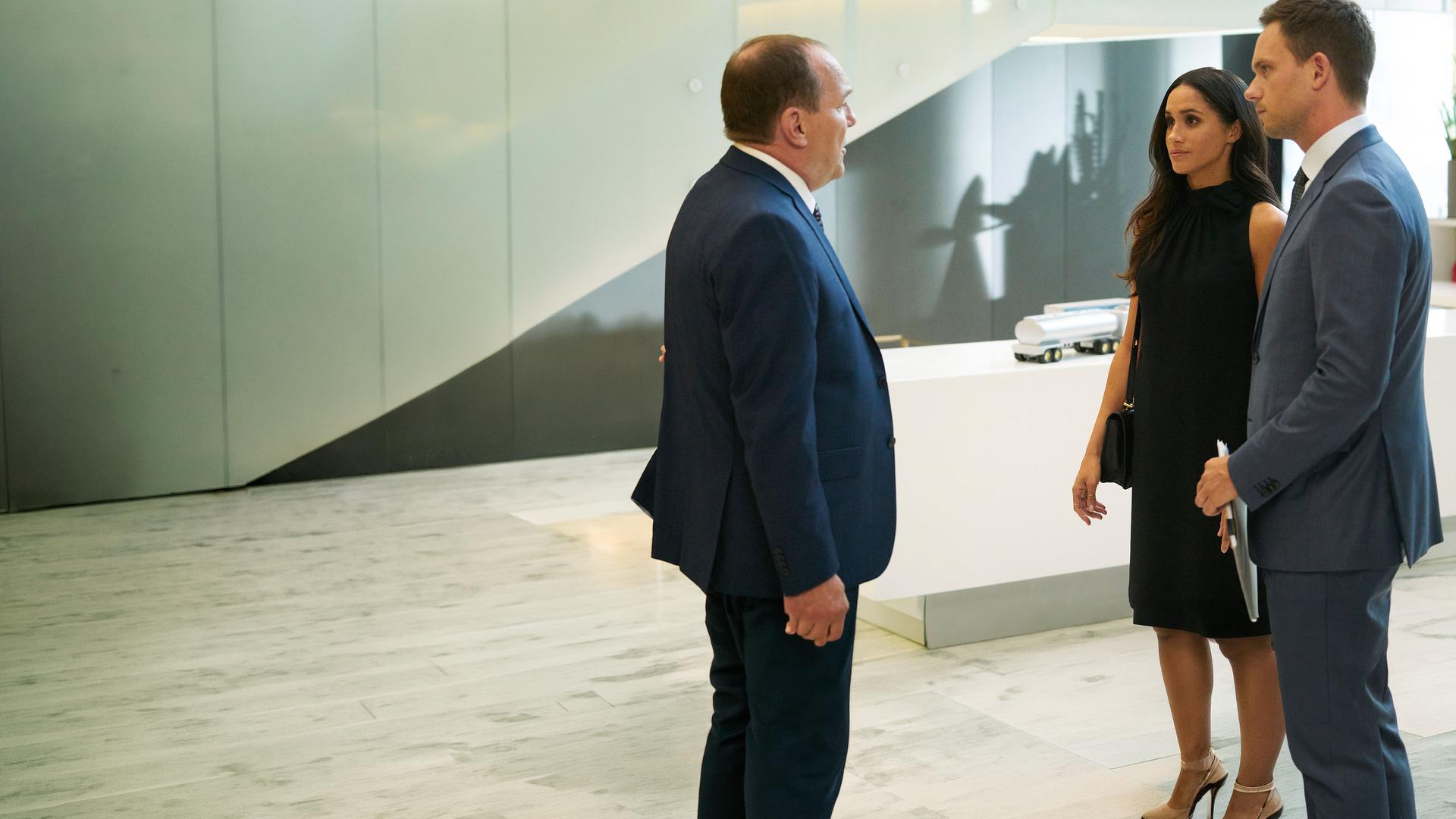 "Suits - Zerwürfnis": Die in schwarz gekleidete Rachel (Meghan Markle) und Mike (Patrick J. Adams), der in seinen Händen ein Dokument hält,  hören konzentriert Larry Burnham (Larry Day) zu, der ihnen gegenübersteht.