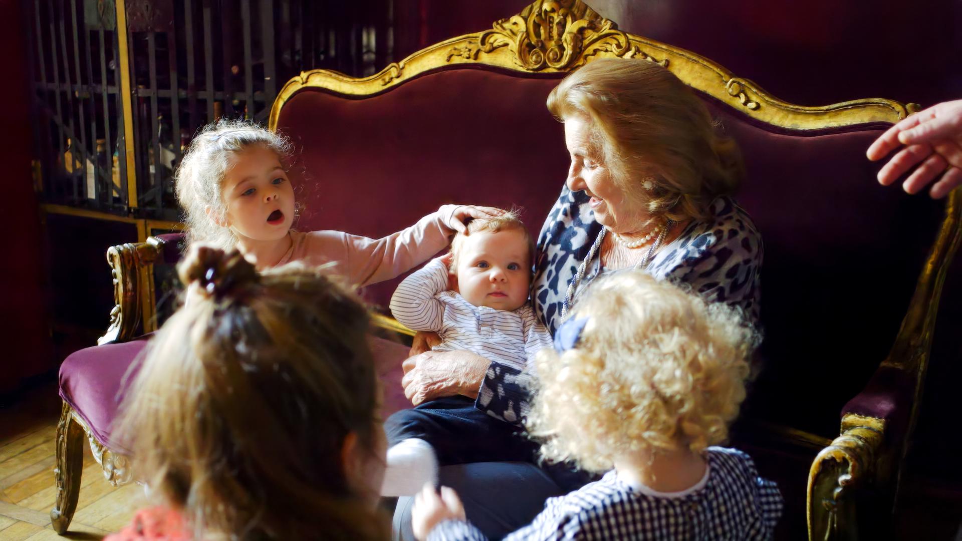 Auf dem Bild sieht man eine sehr alte Frau auf einen edlen, antiken Sofa mit Goldrand sitzen. Sie hält ein Kleinkind auf dem Arm, neben ihr sitzt ein etwa 3-jähiges Mädchen, das dem Kleinkind über den Kopf streichelt. Die alte Frau trägt feine Kleidung und Ohrringe und lächelt das Kleinkind an. Vor dem Sofa sieht man ein weiteres, blondgelocktes Kind von hinten, daneben, ebenfalls von hinten eine Frau mit braunen Haaren und Dutt.