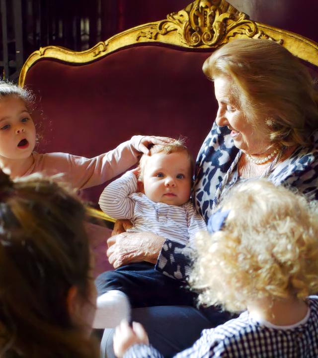 Auf dem Bild sieht man eine sehr alte Frau auf einen edlen, antiken Sofa mit Goldrand sitzen. Sie hält ein Kleinkind auf dem Arm, neben ihr sitzt ein etwa 3-jähiges Mädchen, das dem Kleinkind über den Kopf streichelt. Die alte Frau trägt feine Kleidung und Ohrringe und lächelt das Kleinkind an. Vor dem Sofa sieht man ein weiteres, blondgelocktes Kind von hinten, daneben, ebenfalls von hinten eine Frau mit braunen Haaren und Dutt.