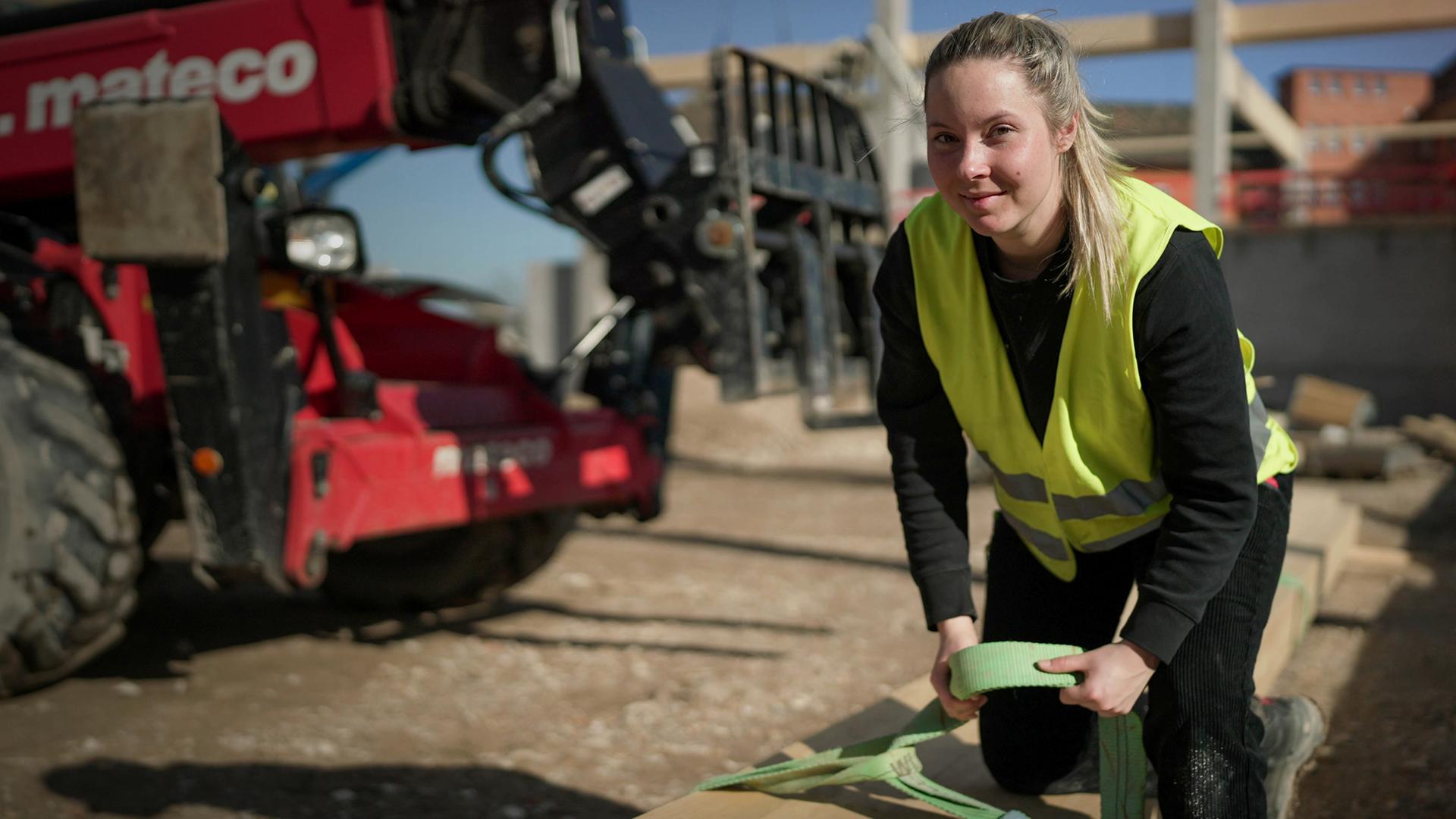 Eine Frau in gelber Sicherheitsweste beugt sich über einen langen Holzbalken und hat einen Gurt in der Hand. Im Hintergrund ist eine Baustelle zu sehen.